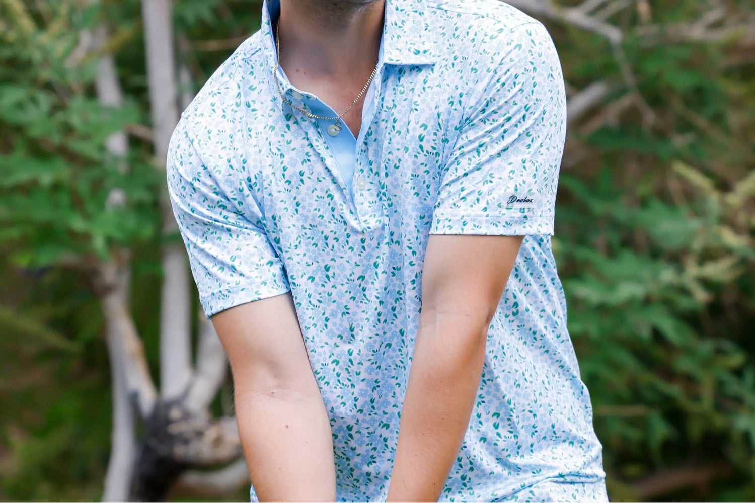 Person wearing a light blue and green floral pattern printed polo shirt outdoors with greenery in the background
